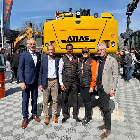 Gruppenbild mit fünf Personen zur Partnerschaft von NAF und ATLAS vor Bagger